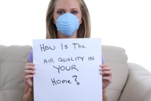 Woman In Mask Holding Sign That Says: How Is The Air Quality In Your Home